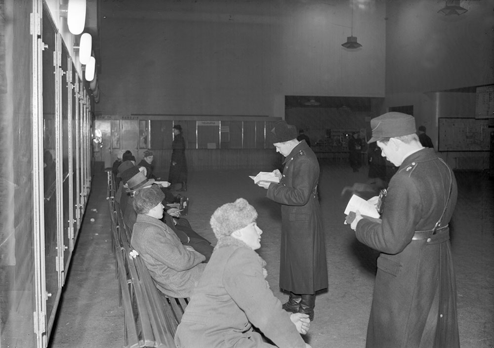 Den militära mössa som kallades "blodskopan" blev en del av polisuniformen 1945. Mössan var mörkblå till färgen, och kanterna som följde sömmarna hade samma färg som kragspeglarna. Till en början användes denna militära mössa endast på vintern. Inspektion av resetillstånd på Tammerfors busstation. Fotograf: E.M. Staf. Bild: Museicentret Vapriikki.