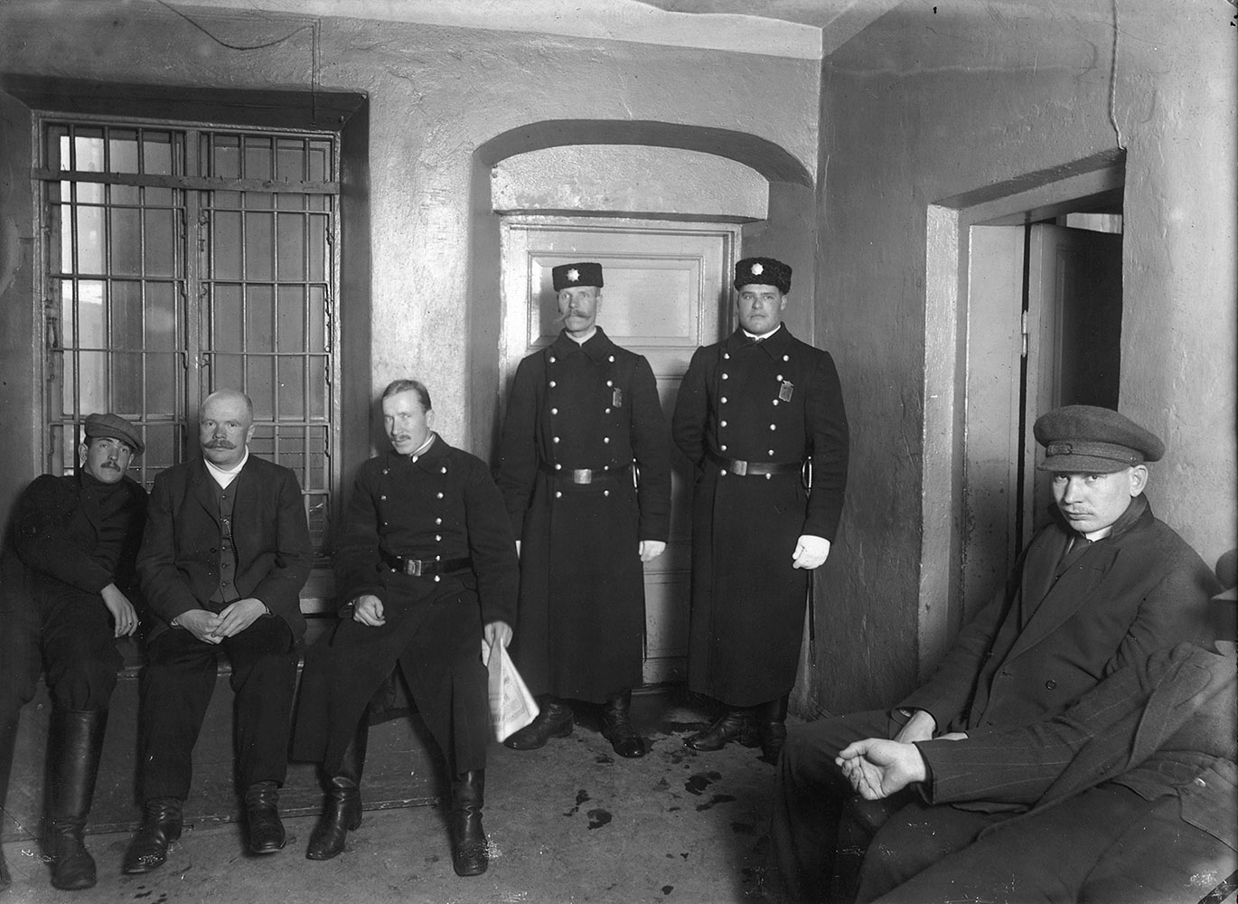 The police overcoats of the early 20th century were military in style, and in Finland they were called "mantteli" (from the German Mantel, overcoat) and "sinelli". The coats were usually made of thick woollen fabric and had buttons in two rows. The colour varied between dark grey and dark blue depending on the uniform statute. Constables and arrestees at a Helsinki jail, around 1910.